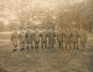 A photograph of ten men posting for a photograph in a grass field surrounded by trees. They are ...