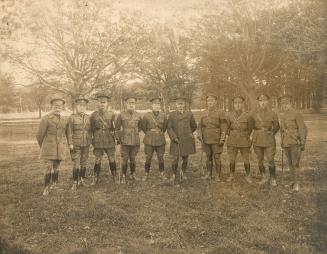 A photograph of ten men posting for a photograph in a grass field surrounded by trees. They are…