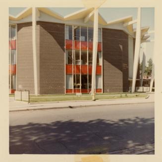 A photograph of a public school, with a paved city street in front of it. There are shadows fro ...