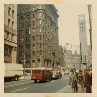 A photograph of a city street, with tall stone buildings on the left side of the photo and a bu ...