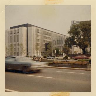A photograph of a large brutalist building beside a wide, paved city street. There is a large t ...