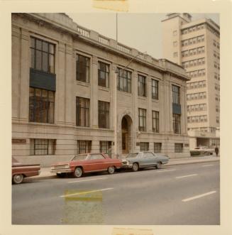 A photograph of a large three story stone building with a paved city street in front of it. The ...