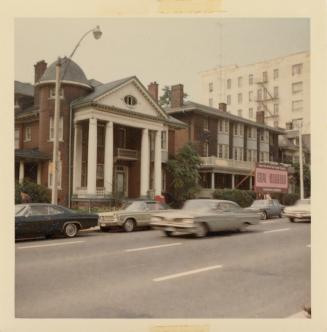 A photograph of a paved city street, with cars parked and driving past a line of buildings. The ...