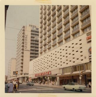 A photograph of a downtown city street, with a paved road in front of a line of skyscrapers and…