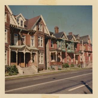 A photograph of a a row of three story residential houses, with a paved city street and sidewal ...