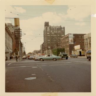 A photograph of a wide city intersection, with a public transit bus, cars and trucks parked and ...