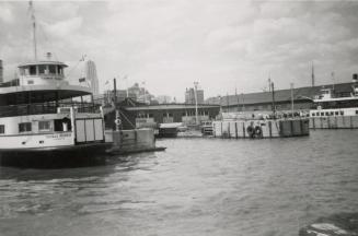 A photograph of a harbour, with docks and a ferry on the left side of the photo. The ferry has  ...