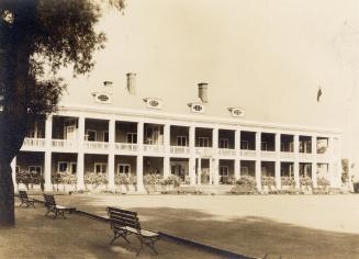 A photograph of a clubhouse with a lawn bowling field in front of it. There is a tree on the le ...