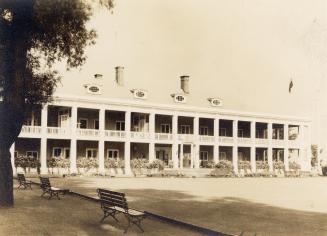 A photograph of a clubhouse with a lawn bowling field in front of it. There is a tree on the le…