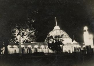 A photograph of a building with a glass dome, lit up at night. There are trees in front of the  ...