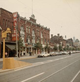 A photograph of a row of brick buildings beside a paved city street. There are cars parked on t ...