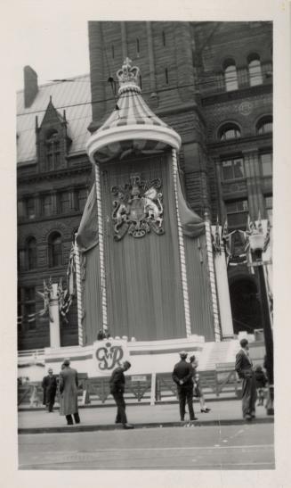 A photograph of a stage erected in front of a large brick building. The stand is decorated with ...