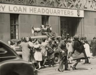 A photograph of a few dozen people sitting and standing on and around a paved city street with …