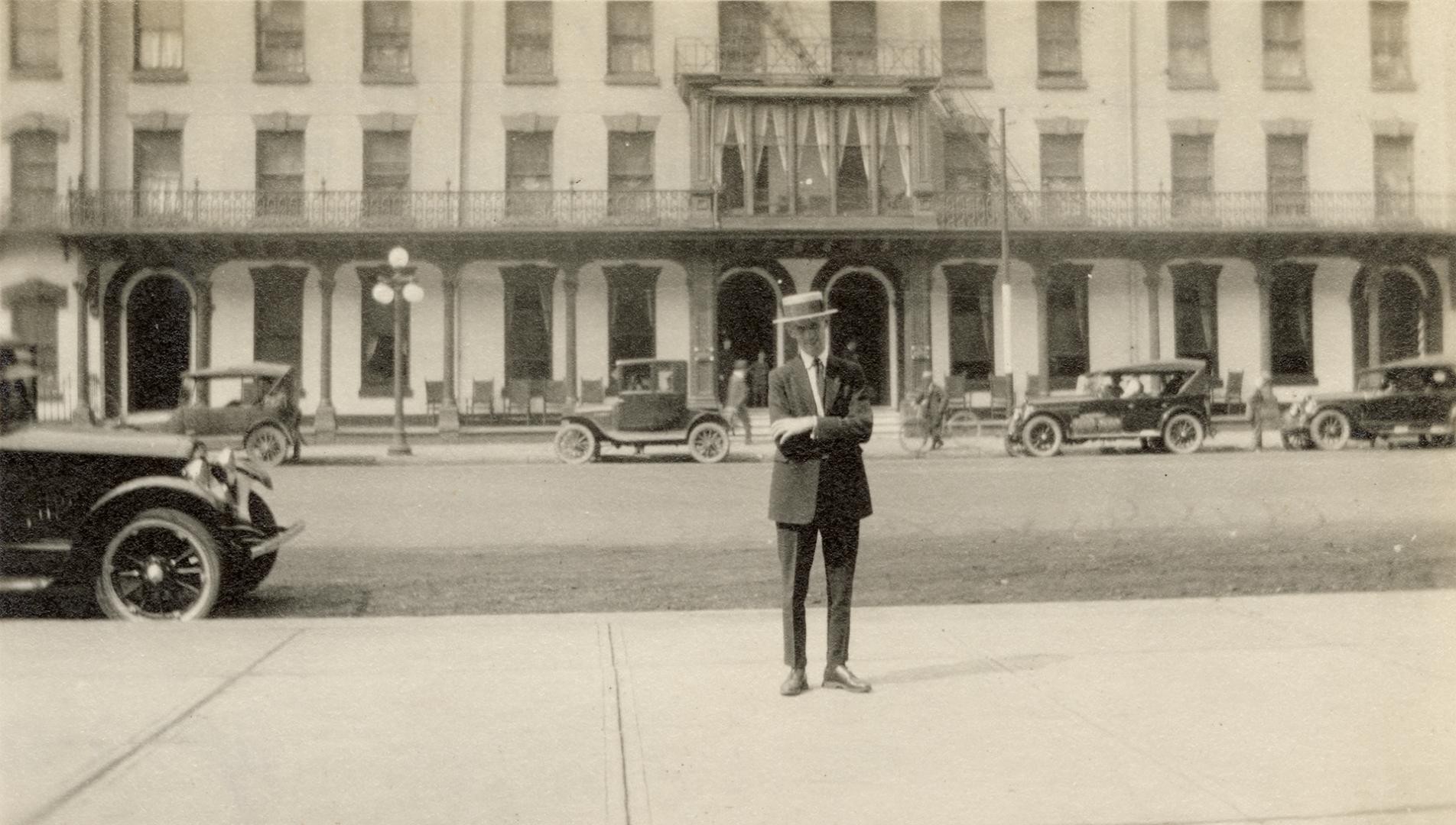 A photograph of a person standing on a sidewalk, with a hotel behind him on the other side of a ...