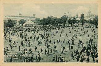 Sepia toned photograph of a vast crowd of people walking on a lawn. Large buildings are in the  ...