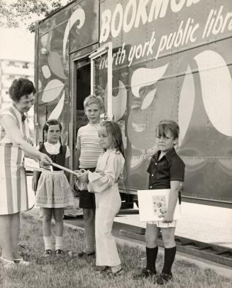 Picture of female librarian, standing in front of a bookmobile, handing a books to a child with ...
