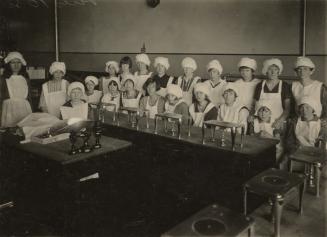 Class picture of girls wearing white hats and aprons at tables with equipment. 