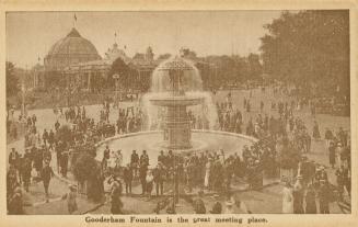 Sepia toned photograph of crowds of people standing around a huge fountain. Large buildings are ...