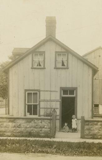 Black and white photograph of two children standing in the doorway of a two story, frame house  ...