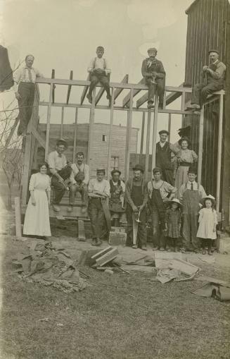 Black and white photograph of men, women and children posing in front of the frame of a house u ...