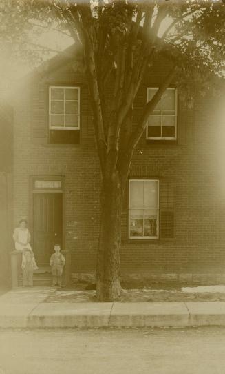 Black and white photograph of a woman with two young children standing in front of a two story  ...