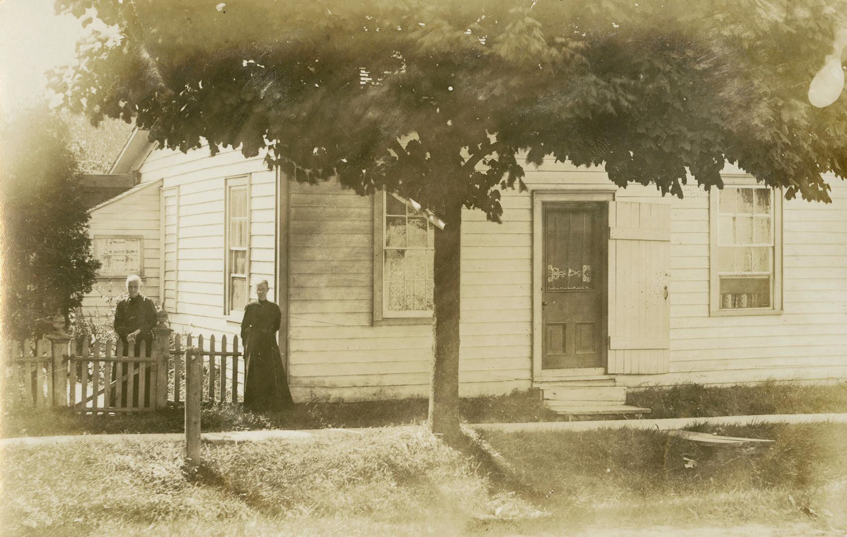 Black and white photograph of two women standing in front of a small, frame bungalow.