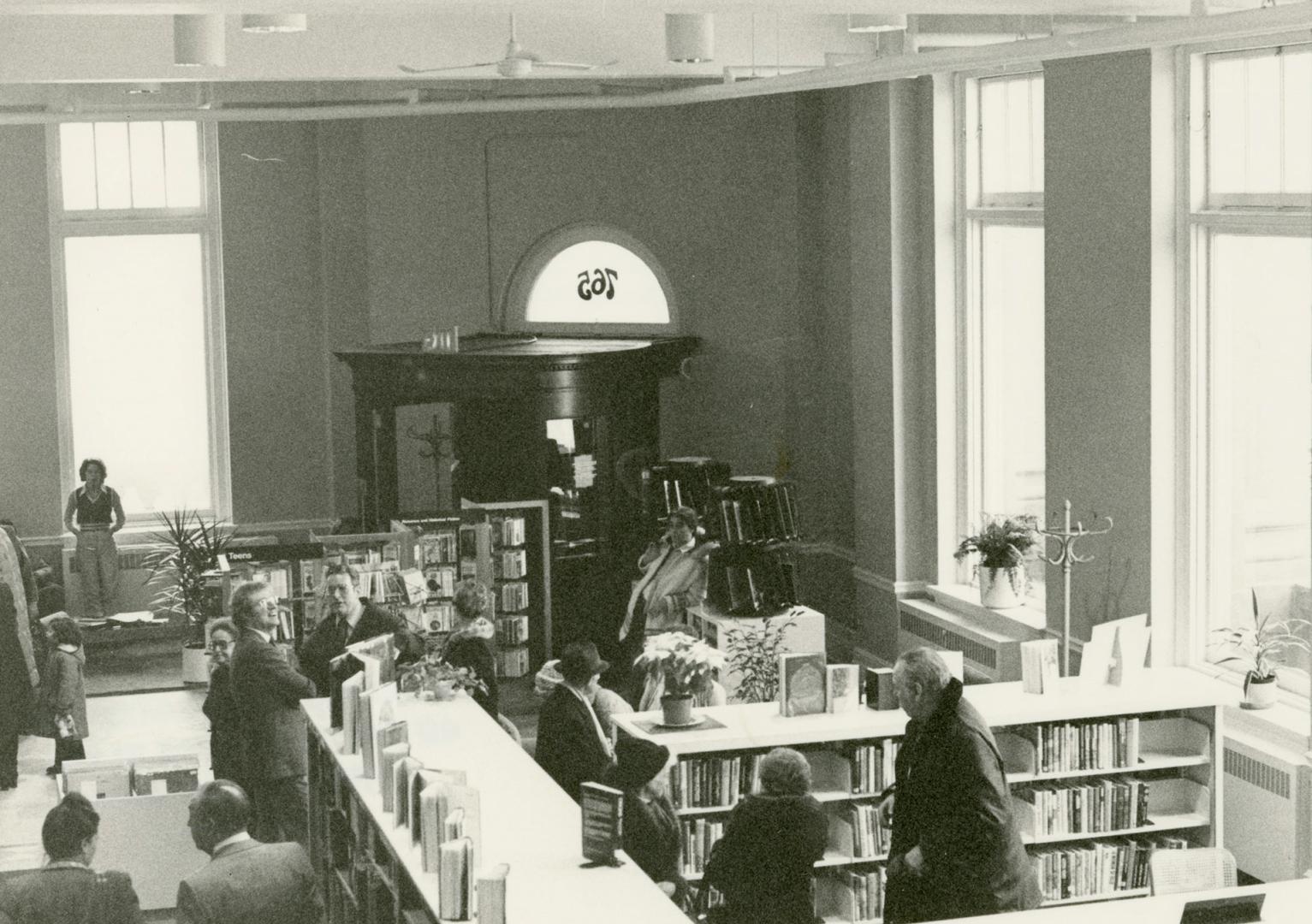 Picture of interior view of library branch showing crowd of people standing amongst shelves. 