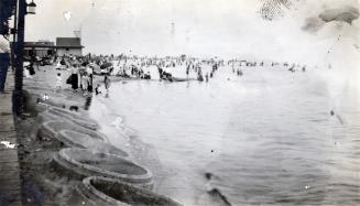 A photograph of a beach, with many people standing and sitting on the beach and swimming in the ...