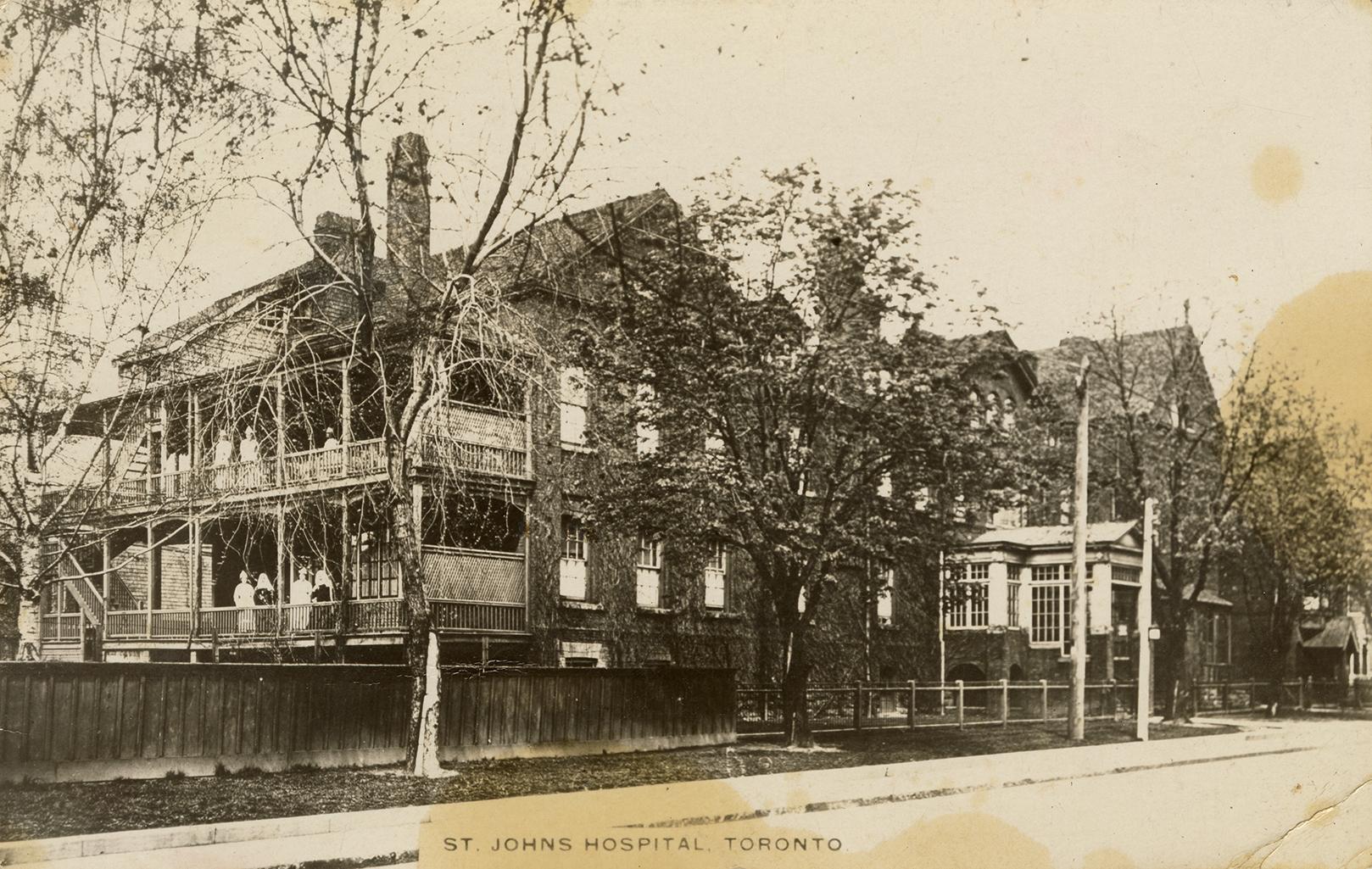 Black and white photograph of a large two story brick house with people standing on wooden balc…