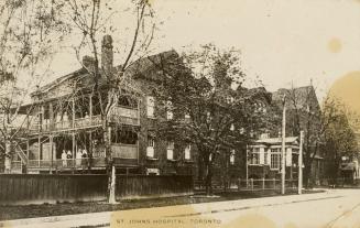 Black and white photograph of a large two story brick house with people standing on wooden balc…