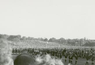 A photograph of a large crowd gathered in a park. There is a hill in the background and a flat  ...