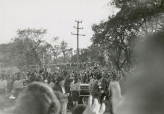 A photograph of a large crowd gathered in a park, standing around a car. There are trees and a  ...