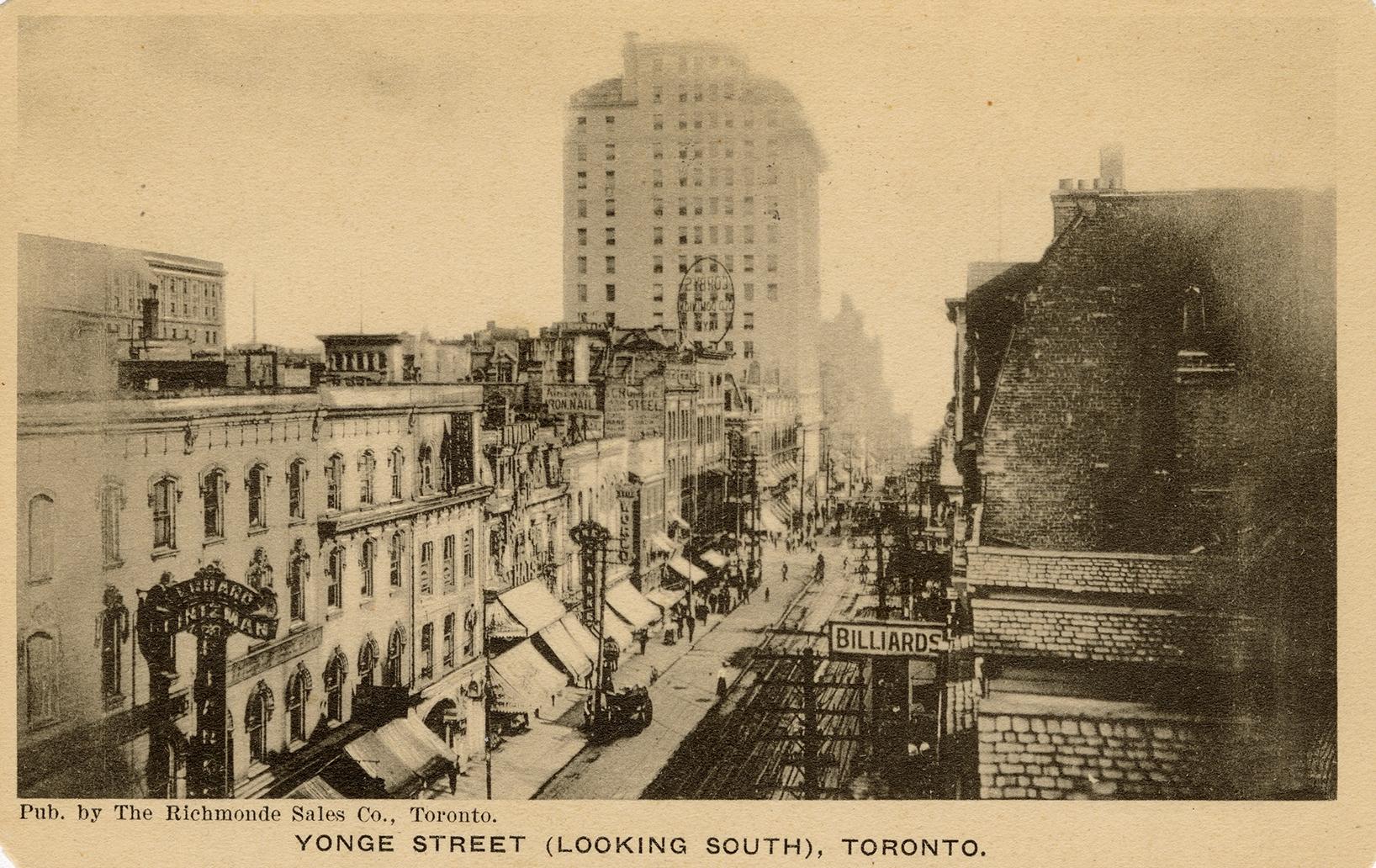 Black and white photograph of very busy city street with tall buildings on either side. Taken f…