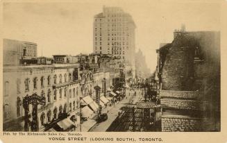 Black and white photograph of very busy city street with tall buildings on either side. Taken f…