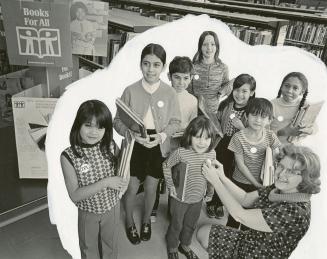 Picture of a group of children in a library and a librarian pinning a button on one child's shi ...