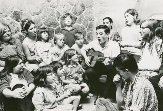 Picture of man playing a guitar surrounded by a group of children singing along. 