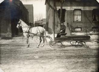 A photograph of a horse hooked up to a sleigh and standing on a city street. There is a man sit…