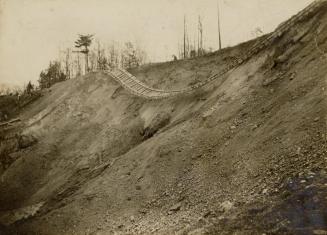 A photograph of a set of train tracks on top of and hanging down over the side of a dirt hill.  ...