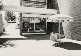 Picture of woman sitting at a table with patio umbrella in front of building. 