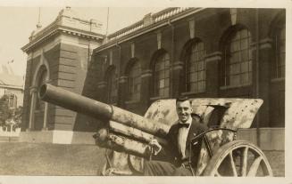 Black and white picture of a man sitting on top of a canon outside of a large building.