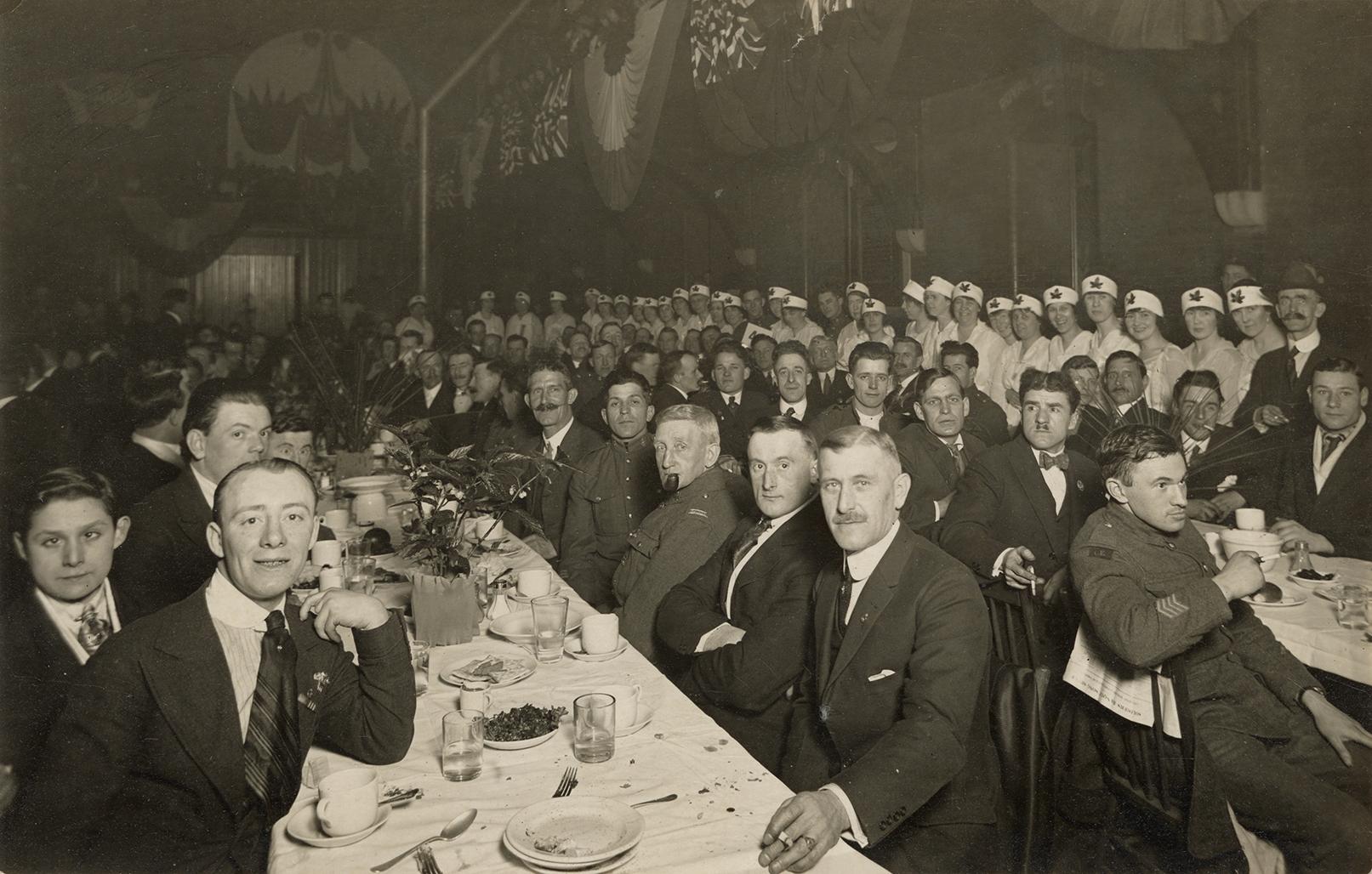 Black and white picture of soldiers sitting in rows of long dining tables. Rows of waitresses i ...
