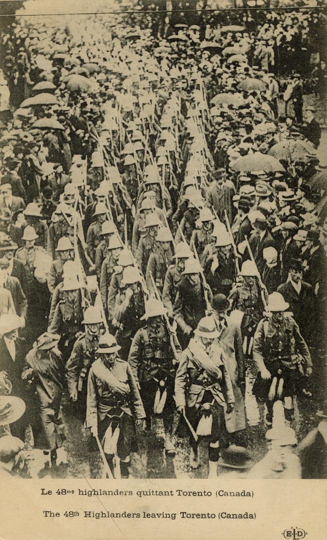 Black and white picture of soldiers in kilts and pith helmets marching down a wide street.