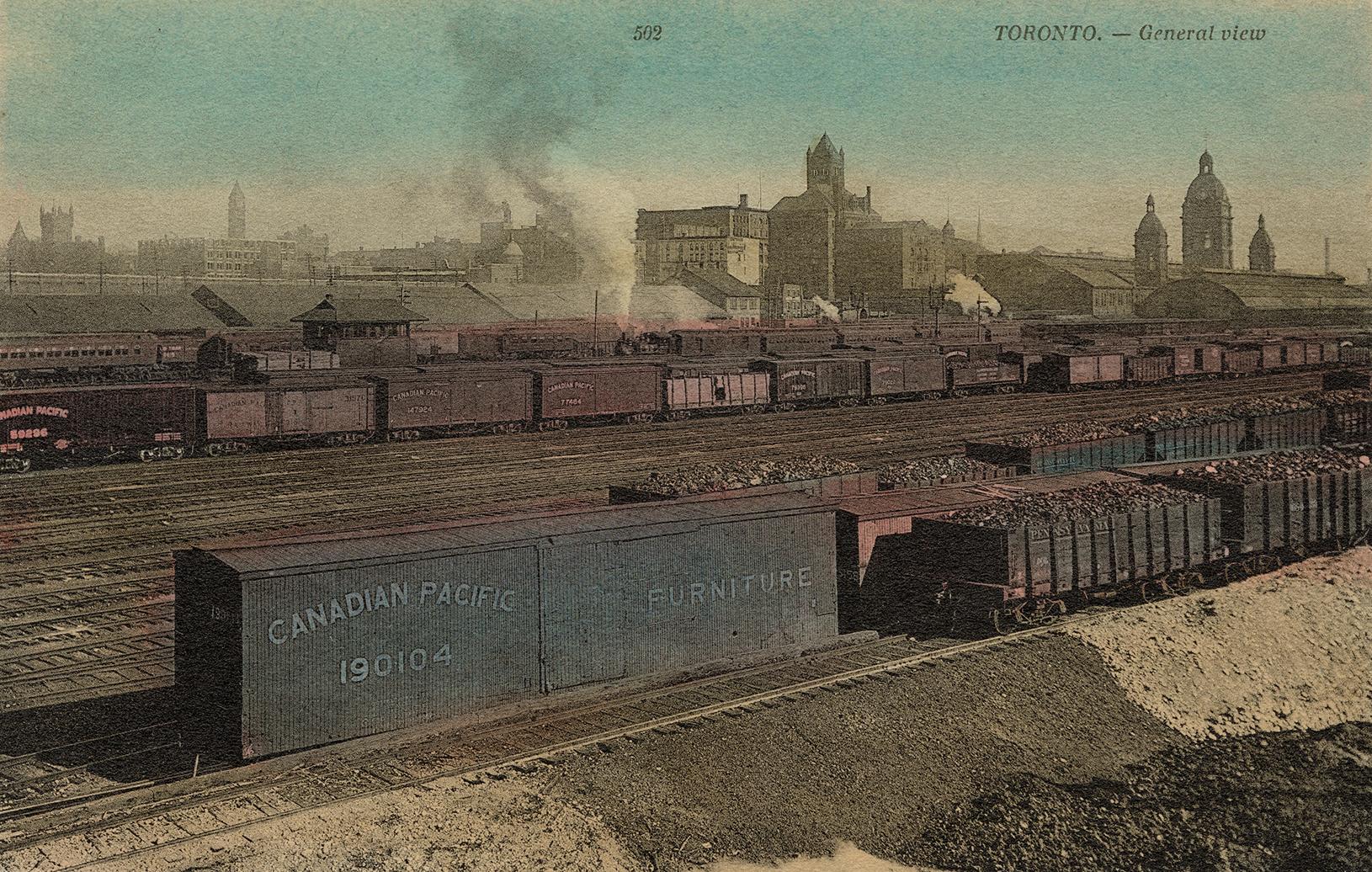 Colour photo postcard depicting a view of railroad cars sitting on many railroad tracks with th ...