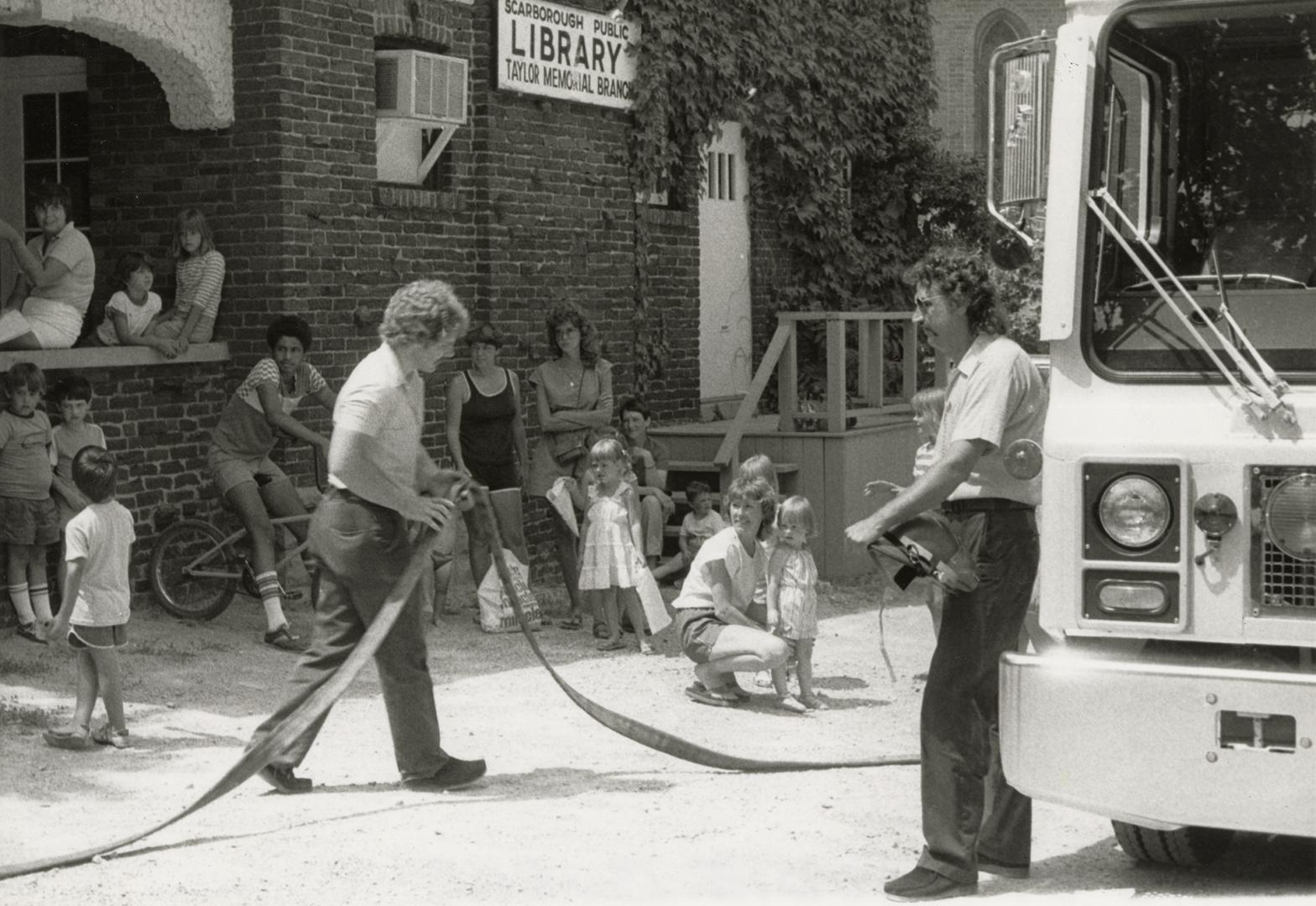 Picture of two fire fighters with hose in front of a crowd of onlookers outside a public librar ...