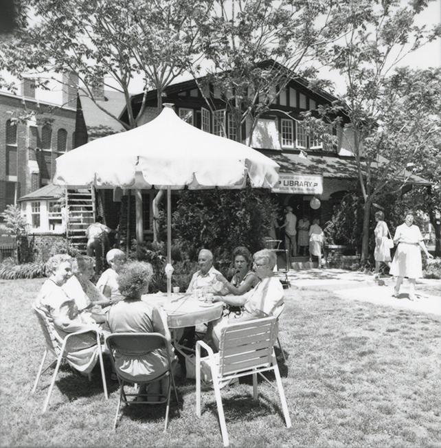 Group of people seated at a table with umbrella at a garden party on library lawn. 