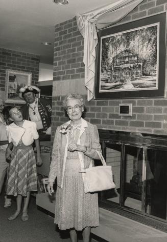 Picture of a woman in front of a fireplace and large painting of library building. 