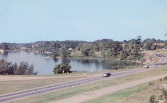 Color photograph of cars driving on a highway beside a body of water.