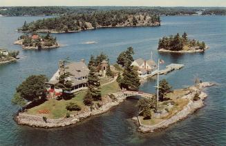 Colorized photograph of a view of a waterway, with a large home on an island connected by a sma ...