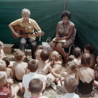 Picture of children sitting outside listening to a librarian storyteller and a guitar player. 