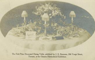 Black and white photo postcard depicting a decorated dining table, adorned with tableware, cand…